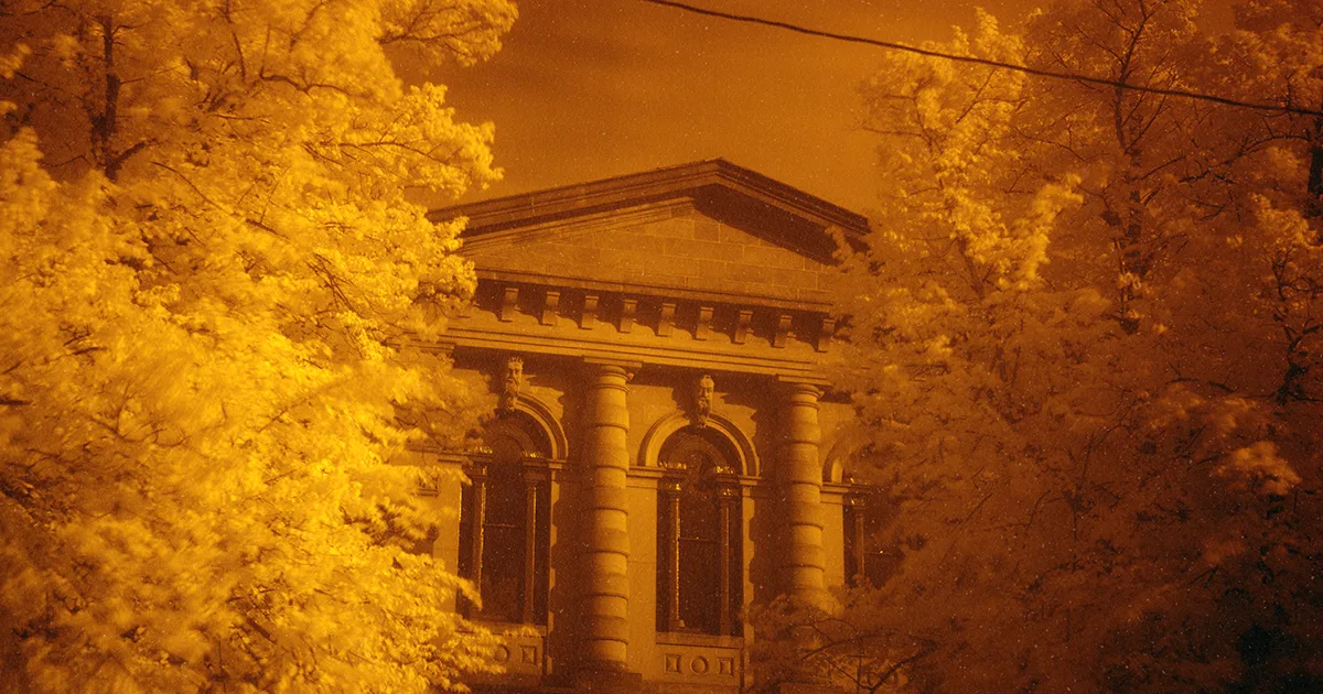 A historic sandstone courthouse with a columned portico and arched windows framed by autumn foliage at dusk